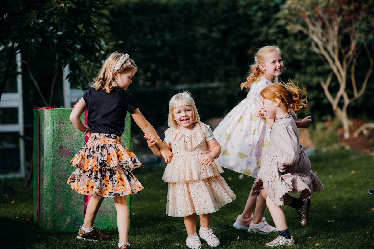 Four joyful children play in a garden, wearing colorful dresses and enjoying a sunny day.